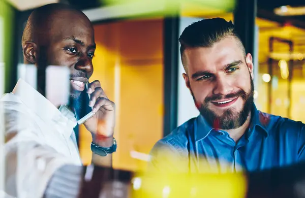 Positive African American executive director calling on smartphone while discussing working plan together with colleague in office behind glass wall with stickers. Partnership in financial company