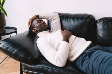 Afro-Amerikan erkek kanepede uzanmış, elleri başının üzerinde ve göğsünün üzerinde kitabı açarken evde yeşil saksıların yanında rahatlarken.