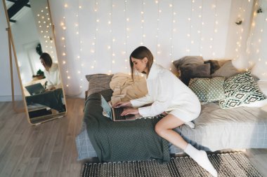 Yatakta oturan ve gündüz vakti evde ışıklarla dekore edilmiş uygulamaları test ederken bilgisayarın ekranına bakan genç kadın yazılım test edicisinin yan görüntüsü