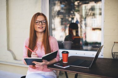 Gülümseyen genç bayan serbest yazar bilgisayarlı akıllı telefon kahve fincanı ile masaya otururken kameraya bakıyor ve pencerenin yakınındaki kafede, bulanık cadde ile birlikte deftere notlar yazıyor.