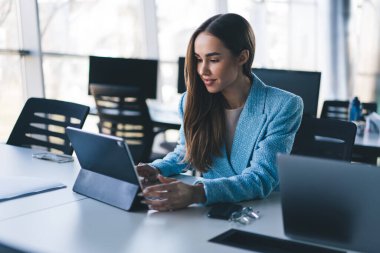 Uzun saçlı, resmi giysiler içinde serbest çalışan bir kadın tablet ve dizüstü bilgisayarla masada otururken kapalı alanda parlak modern çalışma alanı projesinde çalışıyordu.