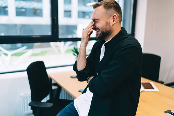 Side view of cheerful bearded male entrepreneur leaning on table and laughing with hand touching nose near window while realizing project mistakes during his stay in office