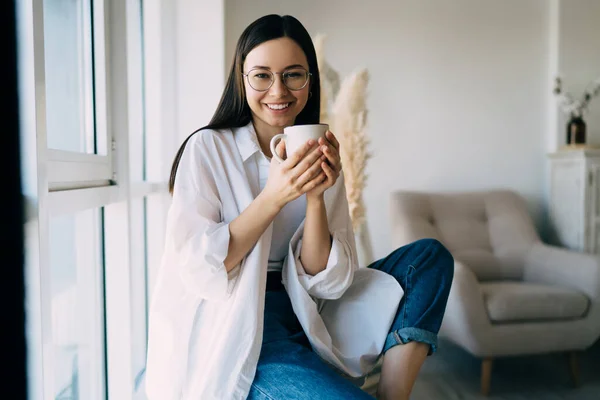 Positive young female in casual clothes looking at camera while smiling and sitting with one leg raised with cup of hot drink in hands in cozy room near glass wall