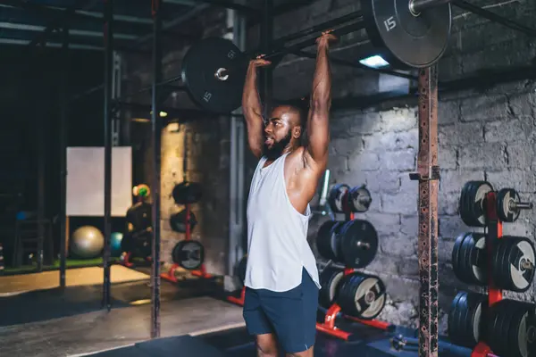 Powerful African American male athlete in white shirt lifting heavy barbell over head while training in gym full of equipment