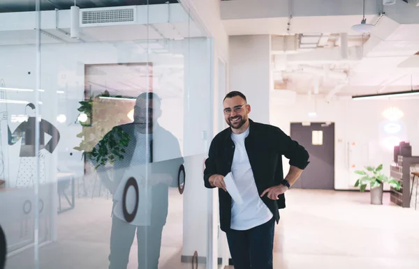 Smiling male entrepreneur standing near reflecting glass wall with hand on waist and looking at camera while moving around neat and clean modern office with documents in hand in free time