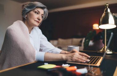Gündelik giysiler içinde, masada dizüstü bilgisayarla oturan ve proje üzerinde çalışırken kameraya bakan olgun kadın serbest yazar.