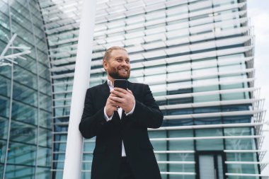 Düşük açılı mutlu sakallı erkek yönetici omuz ile beyaz direğe yaslanıyor ve elinde cep telefonuyla güneş ışığı altında cam duvar binasının girişinde dikilirken diğer tarafa bakıyor.