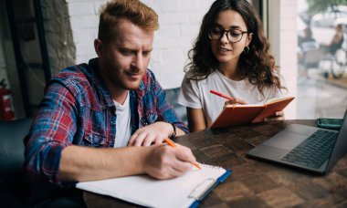 Gözlüklü, dizüstü bilgisayarıyla masada oturan ve kafedeki proje çalışmaları sırasında elinde kalem olan erkek arkadaşının yazılarını okuyan odaklanmış genç bir kadın.
