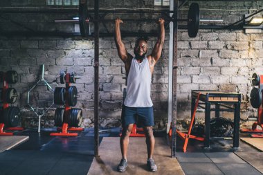 Spor kıyafetlerini giymiş genç Afrikalı kaslı erkek sporcu spor salonunda ağırlıkları kafasının üzerine kaldırarak ağırlık kaldırıyor.