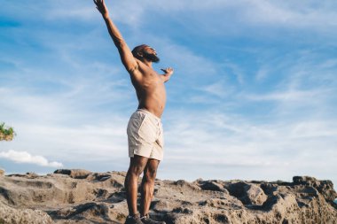 Gömleksiz, şortsuz, kayalık uçurumda duran ve güneşli bir günde bulutlu mavi gökyüzüne karşı doğada tatilin tadını çıkarırken tasasız kollarını kaldıran siyah adamın alçak açılı görüntüsü.