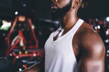 Tarlada tanınmayan genç Afrikalı sakallı, spor giyimli, kaslı erkek sporcunun modern spor salonunda koyu bulanık arka planda duruşu.