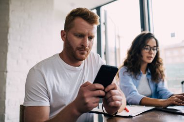 Odaklanmış genç erkek serbest çalışan akıllı telefon ekranına bakarken aynı zamanda dizüstü bilgisayarda pano ile çalışan ve kafede uzak bir projede birlikte çalışan bir kadınla oturuyor.