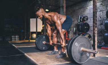 Kararlı genç siyah erkek atlet spor salonunda spor salonunda ağırlık kaldırma egzersizi yaparken arka planda ağırlık kaldırıyor.