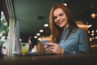 Bilgisayarlı bardak laptopuyla masada oturan, kameraya bakarken gülümseyen ve bulanık kafede cep telefonu tutan genç bir kadın.