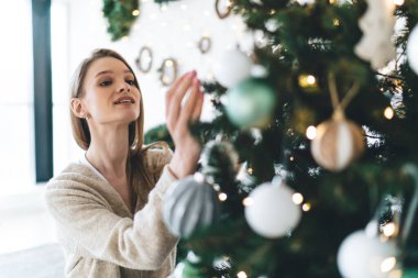 Noel ağacı dalında parıldayan çelenklerle süslenirken rahat bir şekilde gülümseyen genç bir kadının yüksek açısı.