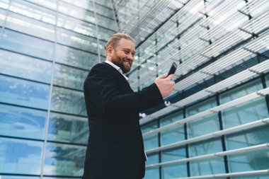 Düşük açılı, gülen iş adamının sokak yansıması ile modern cam duvar binasının yanında duran ve elinde cep telefonu ekranına bakan güpegündüz komik mesajlar okuyan