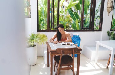 Yukarıdan, akıllı telefon ve gözlüklü, günlük kıyafetli, yaratıcı çalışma alanındaki ahşap masada otururken not defterine not alan genç ve ciddi bir kadın.