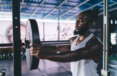 Spor giyim kuşamında güçlü Afrika kökenli Amerikalı erkek sporcunun portresi. Spor salonunda egzersiz yaparken ağırlık kaldırma tekeri bardaydı.