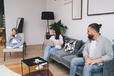 Sakallı baba gündelik kıyafetlerle kanepede oturmuş dizüstü bilgisayar kullanan bir kadınla kız akıllı telefonda video oyunları oynuyor.