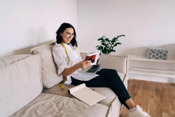 Full body of young female tailor in smart casual wear and glasses sitting on couch with magazine while working remotely on netbook