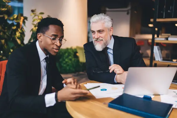 Multiracial colleagues in fancy clothes sitting at wooden desk with papers and computer in modern office while black man looking at cellphone screen in work prosses