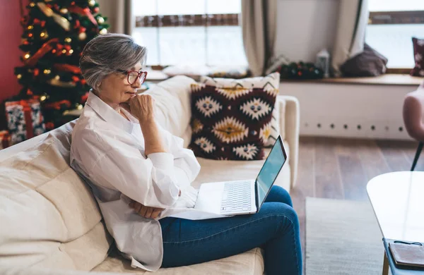 Side view of aged female writer in casual clothes and eyeglasses sitting on sofa in deep imagination with hand at chin and reading draft article before sending to press in living room at home