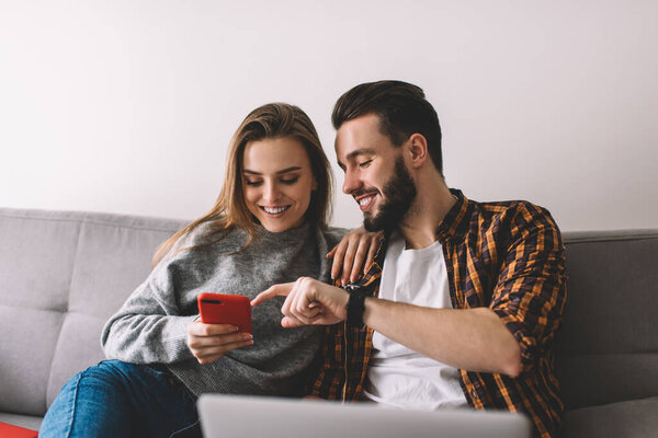 Cheerful couple in casual wear smiling and surfing smartphone together while resting on comfortable couch with laptop during remote work