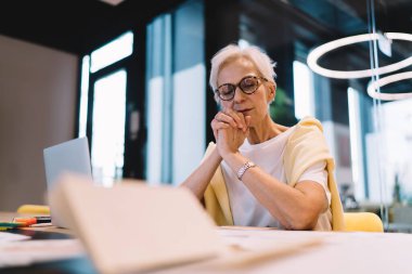 İş yerindeki cam duvarın yanında günlük giysiler ve gözlük takan yaşlı iş kadınının düşük açısı proje üzerinde çalışırken...
