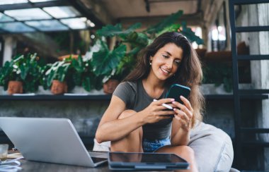 Kalemle gülümseyen ve cep telefonu ekranına bakan genç bir kadın tavan arası kafede dizüstü bilgisayarlı kahve bardağıyla otururken saksıda bitkiler var.