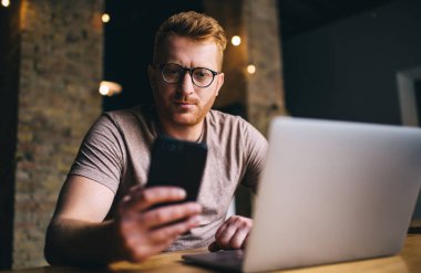 Gözlüklü ve gündelik giyinen konsantre bir erkek serbest yazar kafede oturmuş, ağ kitabıyla projeler üzerinde çalışırken cep telefonu mesajlaşması takıyor.