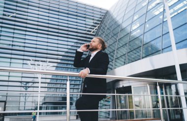 Kendine güvenen genç sakallı iş adamının yan görünüşü. Resmi giyinmiş, korkuluklara yaslanmış, arka planda modern binayla birlikte başka yere bakarken telefon görüşmesi yapıyor.