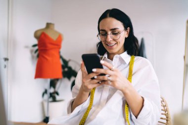 Işık yaratıcı stüdyosunda çalışırken cep telefonuna bakarken gülümseyen gündelik giysiler ve gözlüklerle genç, pozitif moda tasarımcısı.