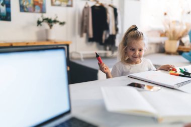 Küçük kız keçeli kalemlerle not defteri çizerken annesi dizüstü bilgisayarla ve planlamacıyla çalışıyor ve ev işi yapıyor.
