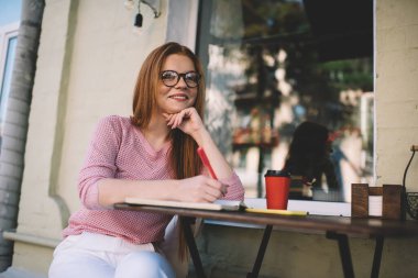 Gözlüklü, gülümseyen ve kameraya bakan genç bir kadın elinde kahve fincanı, elinde akıllı telefonuyla oturmuş günışığında kalemle sokak yansıması ile sokak penceresinin yanında oturuyor.