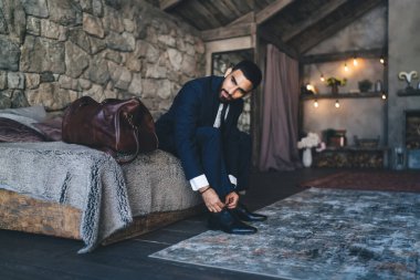 Resmi takım elbiseli etnik sakallı bir adam yatak odasında oturuyor ve bavuluyla uçmayı beklerken bağcıklarını bağlıyor.