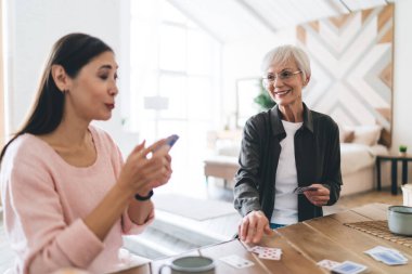 Kağıt oynarken eğlenen kıdemli kız arkadaşlar. Birbirlerine meydan okuyorlar. Açık modern bir dairede oturup sıcak içecekler içiyorlar.