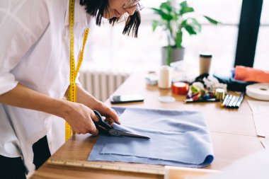 Kumaşı kesmek için makas kullanan isimsiz kadın tasarımcının yan görüntüsü. Akıllı telefon ile ahşap masanın yanında durup kahve fincanı dikiyor ve gündüzleri mağazalarda çalışmaya odaklanıyor.