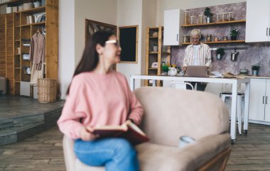 Üst düzey bir kadın mutfakta dizüstü bilgisayar kullanırken kız arkadaşı koltukta oturmuş kitap okuyor ve evde sıcak bir şeyler içiyor.