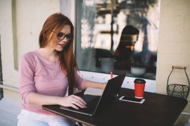 Gündelik kıyafetleri ve gözlükleri olan genç bir bayan bilgisayar ve bir fincan sıcak içecekle masada oturuyor ve uzak bir proje üzerinde çalışıyor.