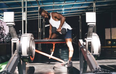 Spor kıyafetli, güçlü sakallı Afrikalı Amerikalı erkek sporcunun kaslı biseps kıvrımlı ağırlık kaldırma hareketini modern spor salonundaki metal barınağın altındaki bankta çalışırken görüntüsü.