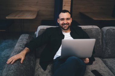 İnternette uzaktan çalışan rahat bir kanepede oturmuş kameraya bakarken gülümseyen ve kameraya bakan sıradan bir adam.