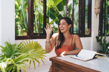 Günlük kıyafetlerle gülümseyen genç bir kadın planlamacı ve kalemle masaya oturuyor ve yaz tatilinde tropikal kafede dinlenirken arkadaşına el sallıyor.