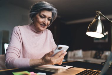 Pozitif orta yaşlı, gri saçlı, sıradan elbiseli bir kadın. Cep telefonuyla sörf yaparken masada dizüstü bilgisayar ve defterle oturuyor.