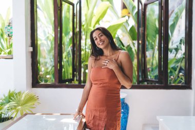 Mutlu gülümseyen, turuncu elbiseli genç bayan gezgin kameraya bakarken parmaklarıyla masaya yaslanıyor ve büyük cam pencerenin yanında elleriyle göğsüne dokunuyor.