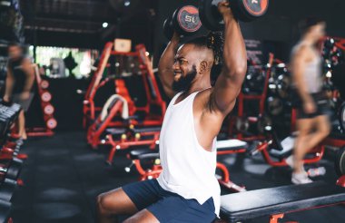Spor kıyafetli Afrikalı sakallı güçlü erkek sporcunun spor salonunda otururken ve spor yaparken kafasının üzerine ağır dambıllar kaldırması bulanık arka planda.