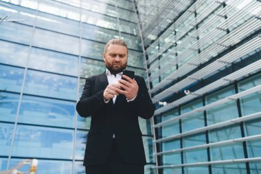 Düşük açılı, düşünceli, sakallı bir işadamı, şehir merkezini yansıtan modern cam duvar binasının yanında dururken elinde cep telefonu ekranına bakıp göz gezdiriyor.