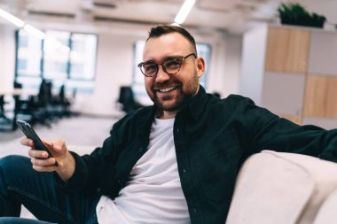 Gözlüklü neşeli sakallı adam modern çalışma odasında koltukta oturuyor ve cep telefonu ve internetten kameraya bakıyor.