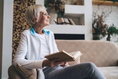 Kitap okurken gülümseyen ve başka tarafa bakan pozitif yaşlı bir kadın hafta sonu evde oturma odasında kanepede oturuyor.