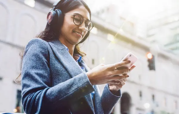 Smiling Indian female listening to music in headphones while standing on city street and relaxing after work. Young woman enjoying playlist in earphones. Watching content in social network, millennial