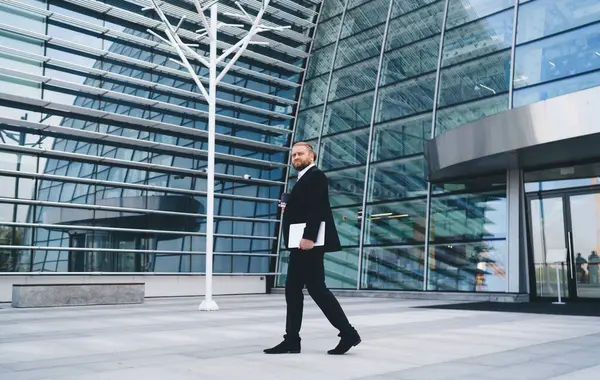 Gündüz vakti modern ofis binasının cam duvarında yürüyen siyah takım elbiseli sakallı işadamının yan görüntüsü.